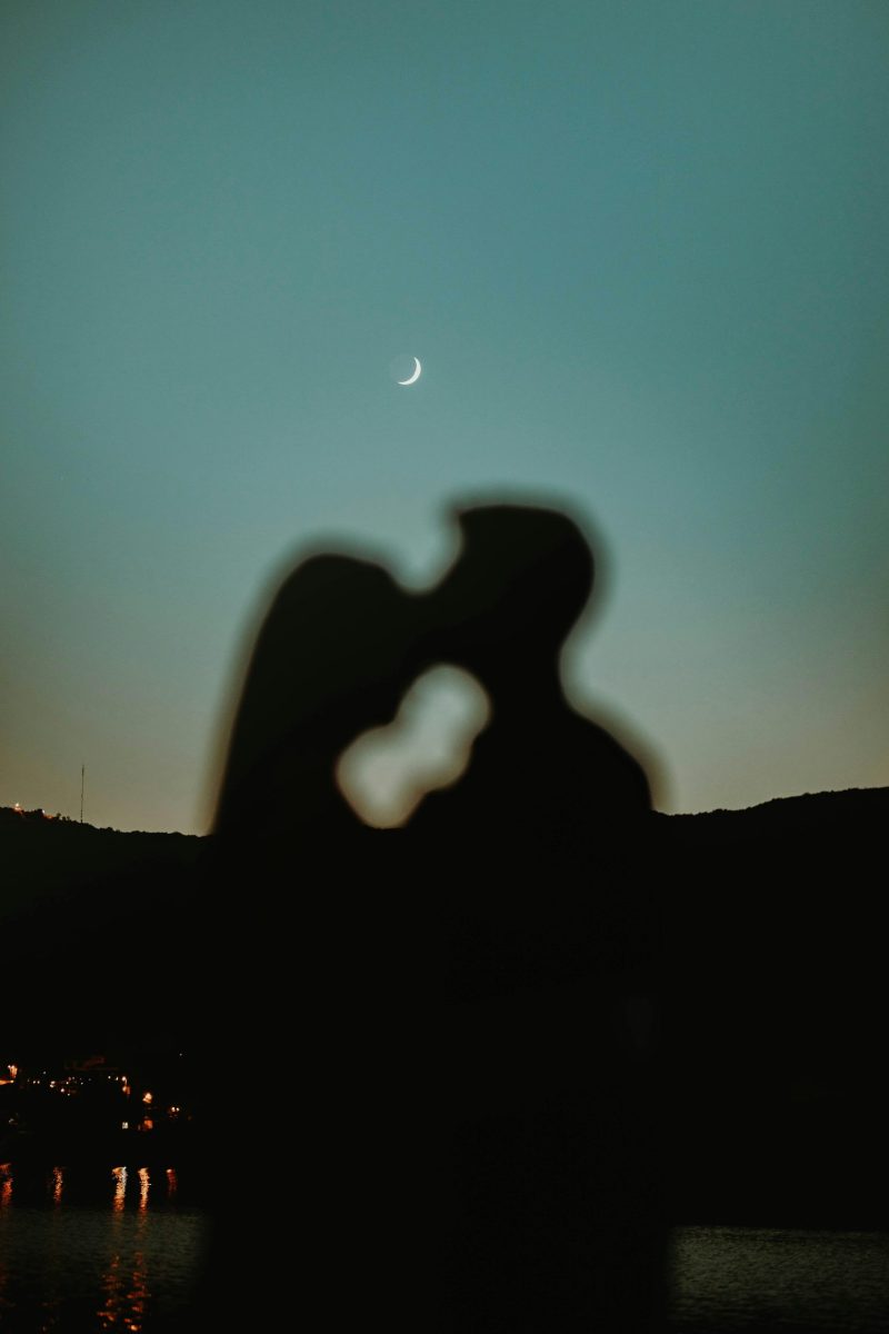 Silhouette of a couple kissing outdoors under a crescent moon at night.